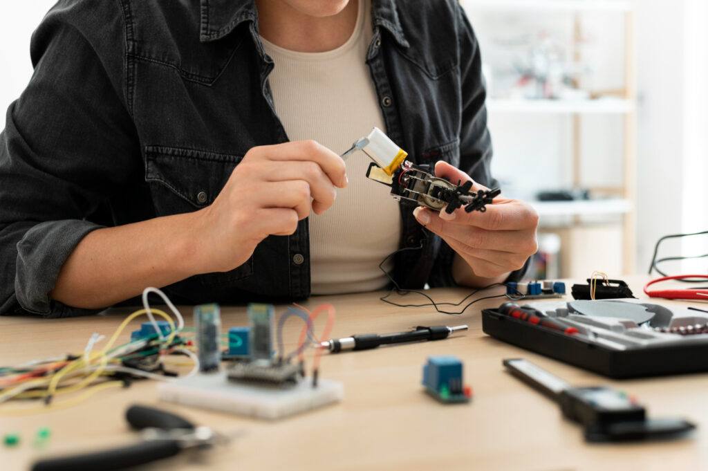 Close-up of a person in a denim shirt working on a small robotic prototype with electronic components, wires, and tools on a wooden workbench.